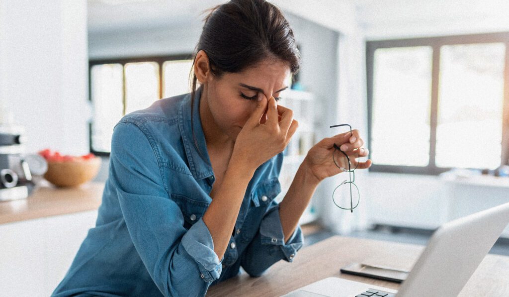 Woman with laptop holding glasses and looking tired which can be a symptom of thyroid issues