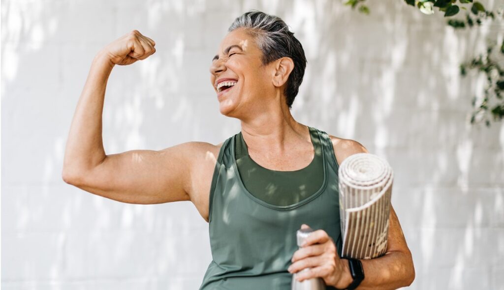 Une femme d'âge moyen tenant une bouteille d'eau et un tapis de yoga célèbre ses progrès en matière de remise en forme en exhibant ses biceps en plein air.