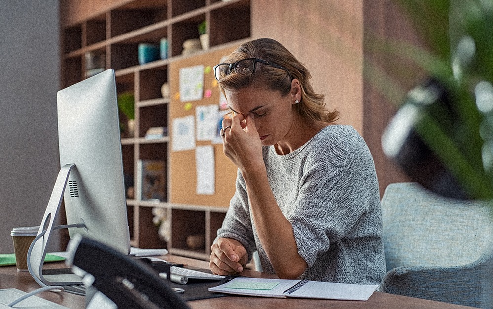 A business woman at her desk with a computer in front of her in an office. She is holding her head to signify a headache, tiredness, and stress.&nbsp;