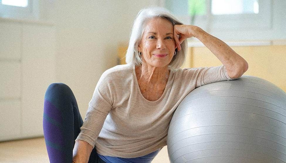 Une femme âgée souriante assise sur un ballon suisse chez elle.