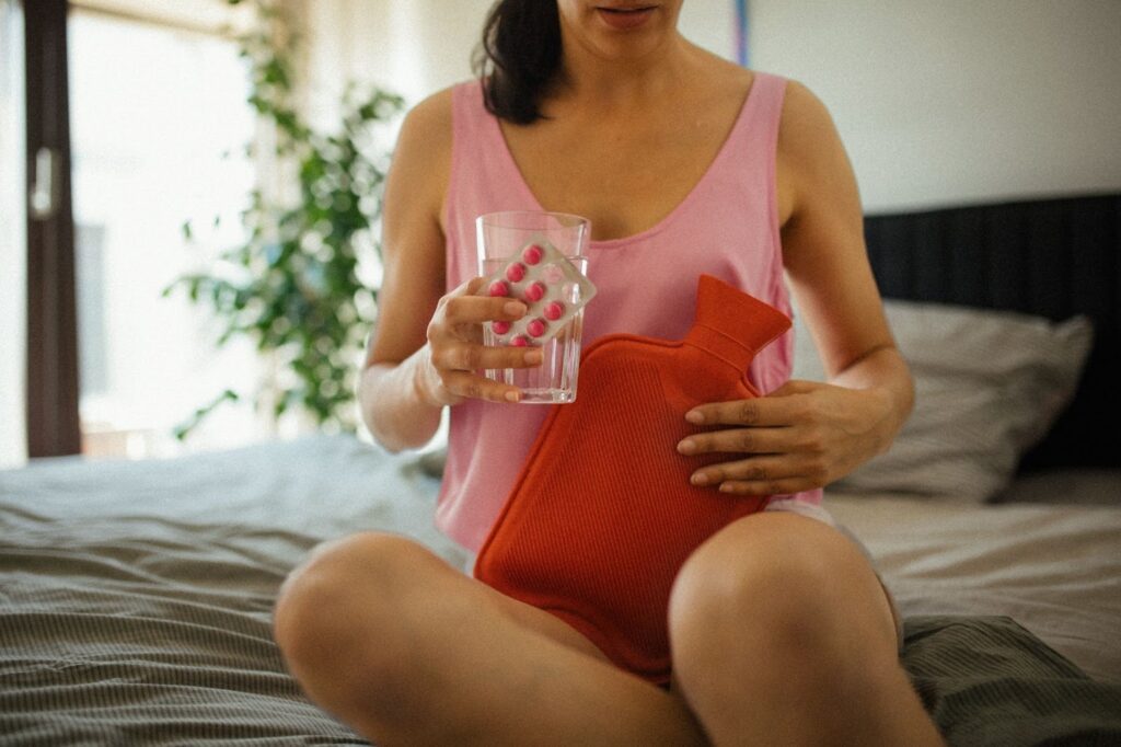 Young woman holding a glass of water, pain medication and a hot water bottle to soothe abdominal and ovarian pain.
